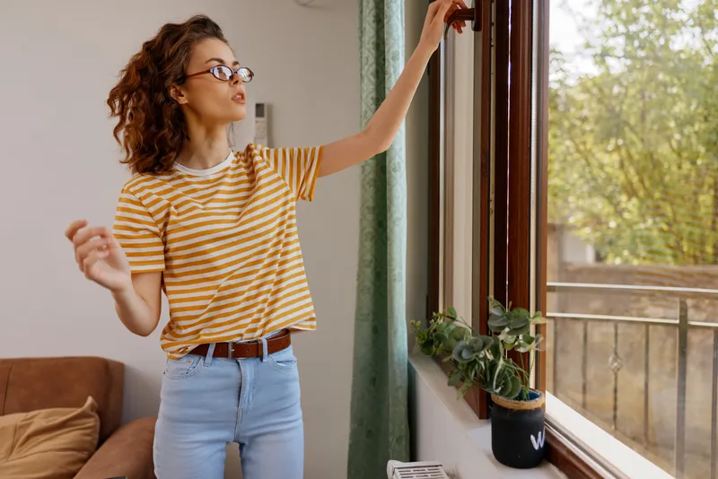 Young woman with curly hair stands by a sunny window, wearing a striped t shirt and jeans. Home lifestyle scene highlighting daily routine and self care in a warm, relaxed mood.