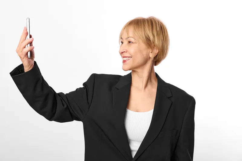 Woman business smartphone smile, pointing gesture in studio white background, mature manager in black blazer portrait, holding phone for selfie, conveying communication technology help