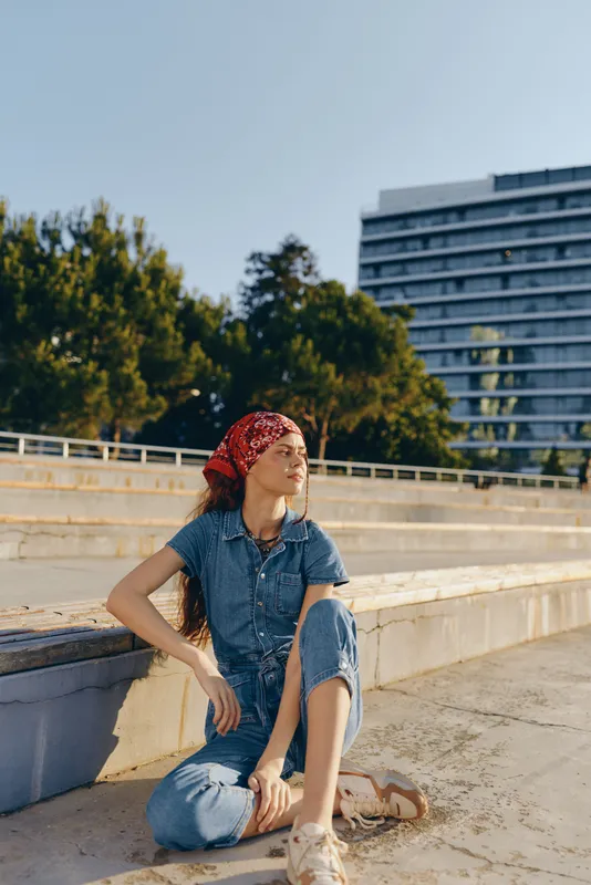 lifestyle woman in modern boho-western denim outfit sitting outdoors with bandana and relaxed street style in warm film color tones