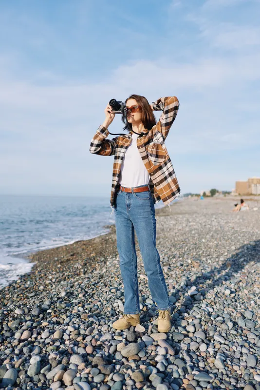 Woman on pebble beach photographed with camera, wearing plaid shirt and jeans, capturing coastal scenery under clear sky, sunlit scene conveying exploration, casual outdoor photography and travel