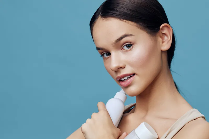 Young model with clean skin holding skincare products, smiling gently against a light blue background, emphasizing a healthy beauty routine and self care