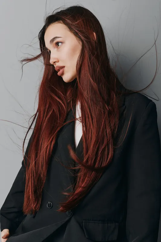 Woman with long red hair wearing black blazer posing against gray wall. Fashion model with natural makeup and serious expression in casual elegant style portrait indoors.