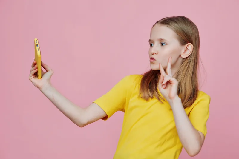 A young girl in a yellow t shirt poses playfully while taking a selfie against a pink background, showcasing joy and confidence in her expression