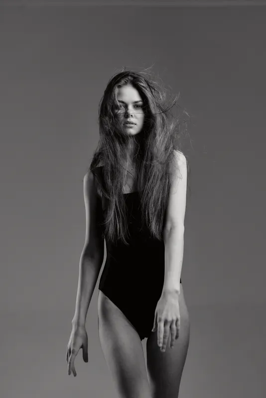 Black and white portrait of a woman with long hair wearing a black bodysuit, expressing calm and elegance with soft studio lighting.