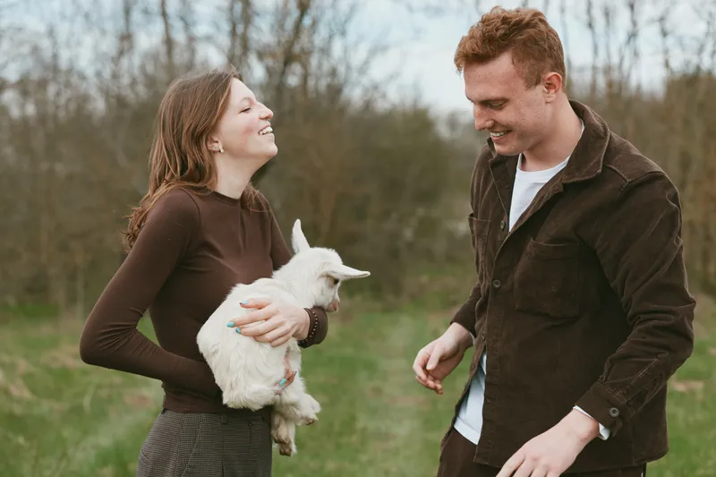 A joyful couple hold a white goat kid in a sunlit field, sharing smiles. A lifestyle moment that highlights companionship, animal bonds, and rural charm in everyday life. Warmth and connection.