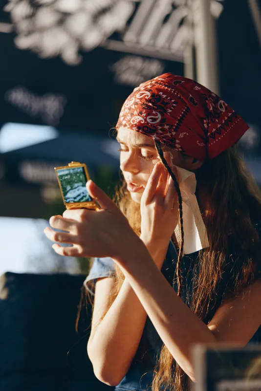 lifestyle woman in modern boho-western denim street style applying makeup using compact mirror with natural light and film color tones, casual urban vibe