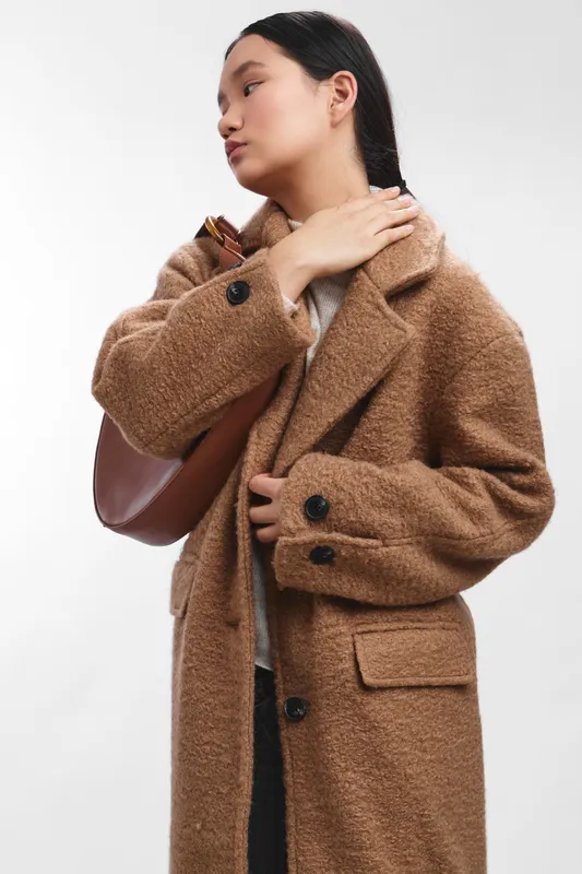 Asian woman wearing a warm brown coat stands against a neutral backdrop, exuding confidence and style as she adjusts her collar in a poised, modern portrait.