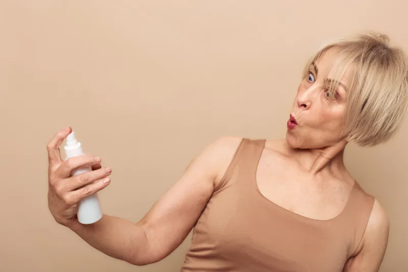 Blonde woman in beige top sprays mist on her face with surprised expression. Skincare routine concept with hydration and beauty treatment on neutral background.