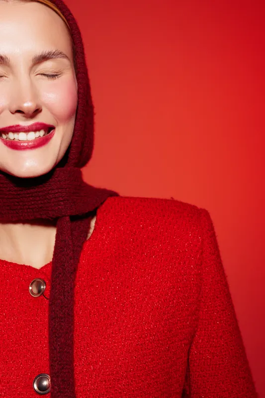 Smiling woman in red coat and scarf with closed eyes stands against red background. Concept of fashion, winter style, happiness, and warm clothing for cold seasons.