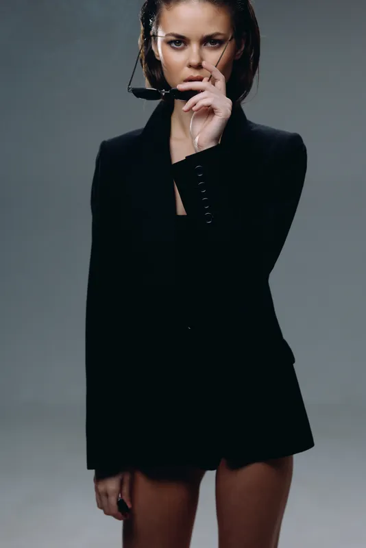 Stylish woman in black blazer holding sunglasses with confident expression and sleek hairstyle on gray background