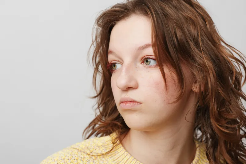 Young woman with red hair and freckles looks away thoughtfully against a plain white background. Concept of contemplation, natural beauty, and casual portrait photography.