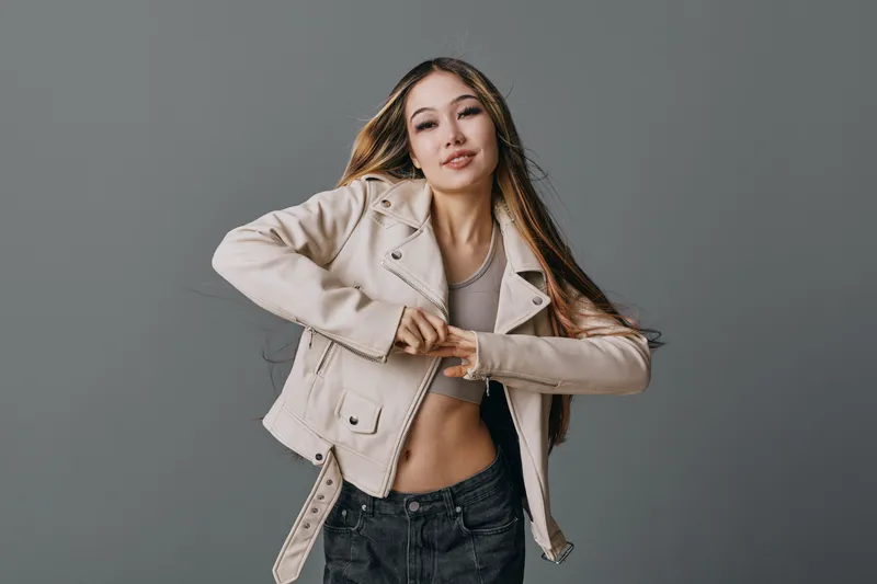 Young woman with long hair wearing beige jacket and crop top posing against grey background. Casual style, confident expression and modern fashion look.