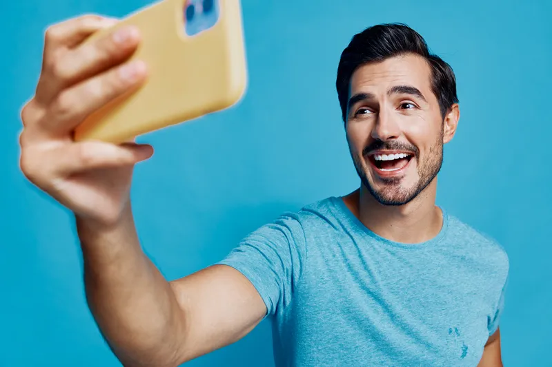 Happy smiling young man taking a selfie with smartphone against bright blue background, conveying a relaxed and joyful mood. People lifestyle concept.