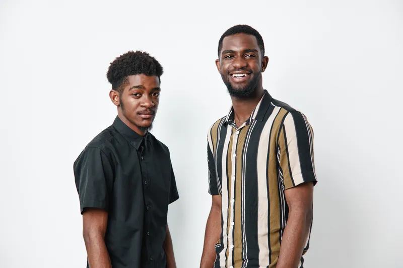 Two young African American men with dark skin standing together against white background, casual clothing, smiling and looking at camera in studio portrait.
