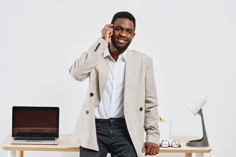 Confident smiling man in beige blazer and white shirt talking on mobile phone and standing at workspace with laptop and desk lamp, white background, studio portrait. Business lifestyle concept
