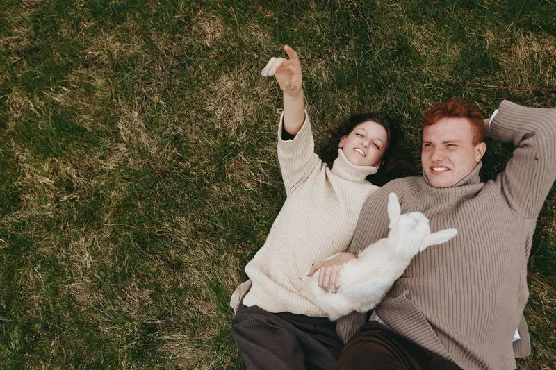 Happy couple relaxing outdoors on grass, taking a selfie with a small white pet, smiling faces, cozy sweaters, casual date, natural light, romantic mood, lifestyle moments, isolated on green lawn.