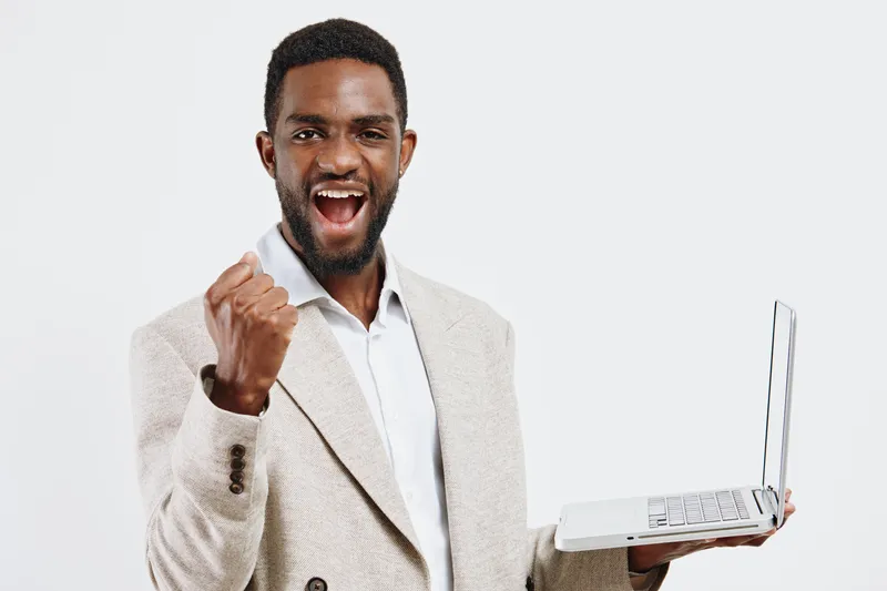 Confident young African American man wearing light coat and white shirt celebrating success with a triumphant fist gesture, holding a notebook in front of plain light gray background, studio shot. Peo