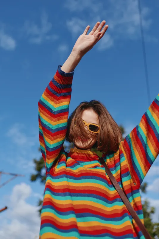 Woman in her twenties wearing a colorful striped sweater and yellow sunglasses, joyful with arms raised outdoors under blue sky, casual outfit for travel and vibrant lifestyle scene.