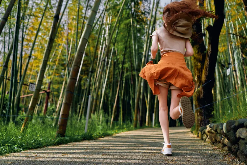 Woman running through bamboo forest on a shaded path wearing an orange dress and white sneakers, back view with hair and skirt in motion, nature trail scene conveying freedom and summer energy and