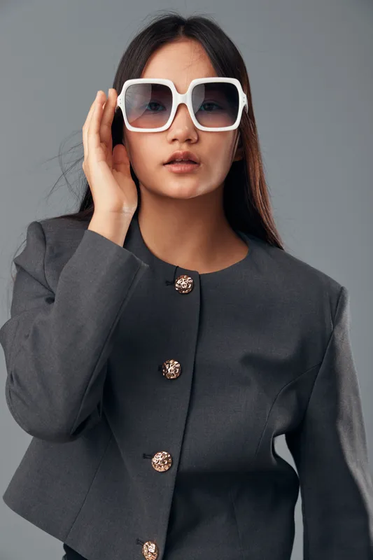 Woman wearing large white sunglasses and gray jacket with decorative buttons looking forward on gray background. Fashion, portrait, style, model, casual, confident, modern, elegance.