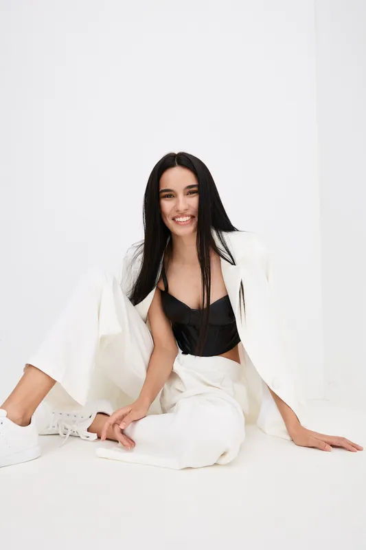 Smiling woman with long black hair wearing white pantsuit and black top sits relaxed on white floor against minimalistic white background, radiating joy and style.