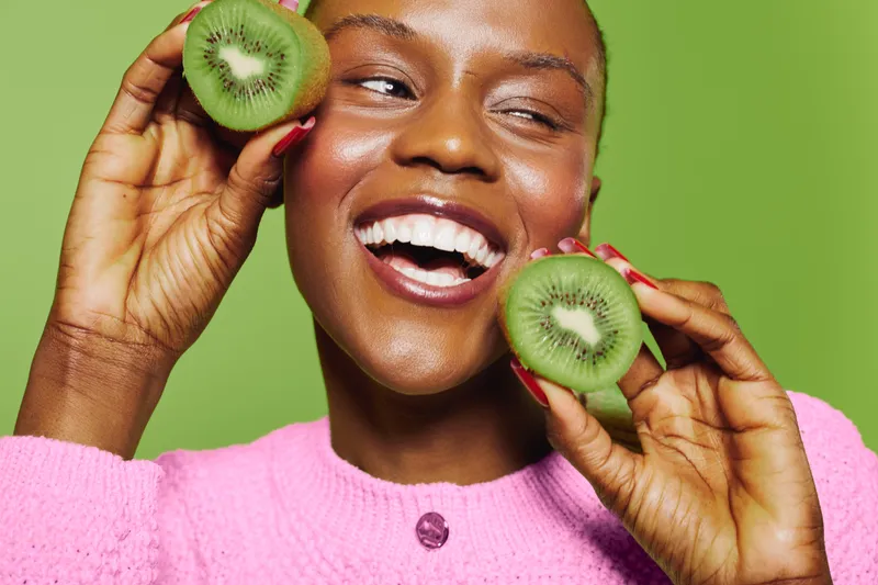 Smiling dark skinned African woman holding fresh kiwi fruit slices near face on green background. Concept of natural beauty, healthy lifestyle, fresh fruit, and joyful wellness.