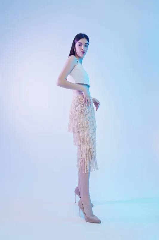 Young woman in a white top and fringe skirt poses confidently in high heels. Studio shot with soft blue lighting. Concept for woman beauty fashion and elegance.