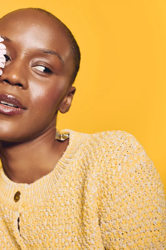 Close-up of a smiling black woman with dark skin wearing a yellow knitted sweater holding white flowers near her face. Bright yellow background creates a warm and cheerful mood.