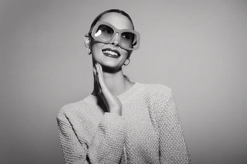Stylish smiling white woman with light skin wearing sunglasses and knit sweater poses against neutral background.