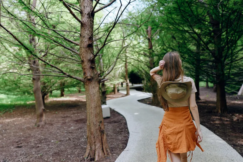 Woman walking on a winding path through a green forest nature trail, wearing a sun hat and orange skirt, casual travel and outdoor exploration with serene woodland atmosphere