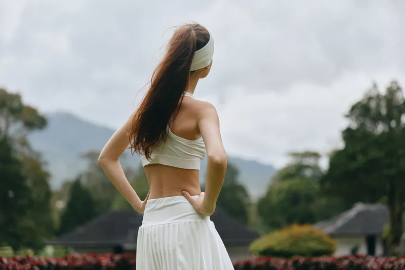 Young woman in sporty attire enjoying nature, standing with hands on hips, showcasing healthy lifestyle and fitness concept in outdoor setting with lush greenery