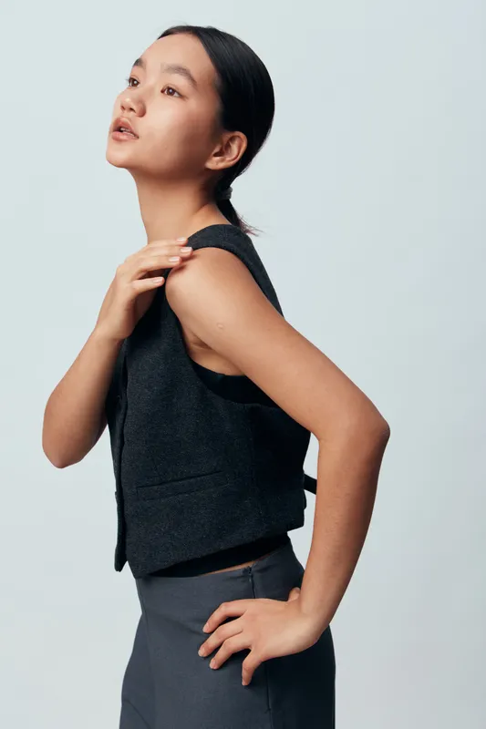 Young woman with dark hair pulled back wearing sleeveless black top and fitted pants, posing against light background with hand on shoulder and side profile expression.