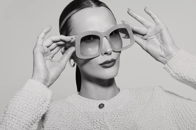 Stylish white woman with light skin wears oversized sunglasses and textured sweater posing confidently in monochrome studio. Concept for fashion, elegance, and accessories.