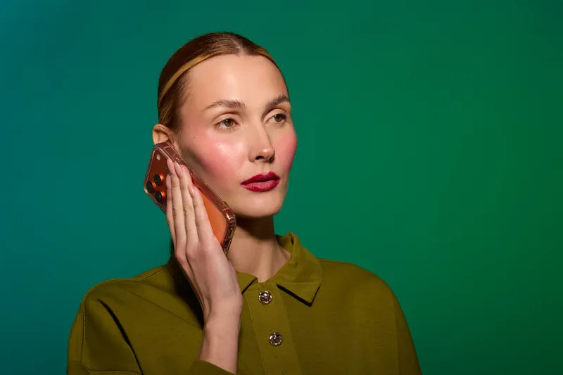 Young woman with clear skin and red lips holds smartphone to ear, looking away on green background. Concept of communication, technology, and modern lifestyle.