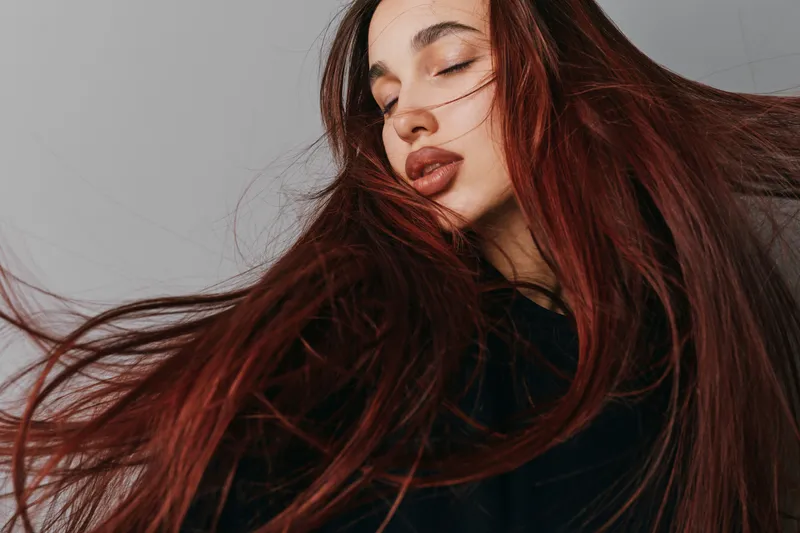 Woman with long red hair and closed eyes posing for portrait with natural makeup and serene expression on gray background. Beauty, fashion and hairstyle concept with flowing hair movement.