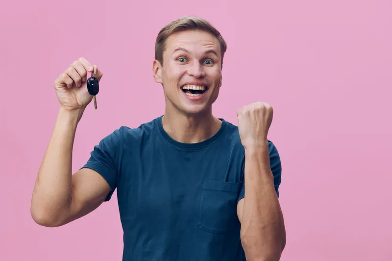 Smiling man with short brown hair wearing casual blue t-shirt holding car key isolated on pink background, celebrating success, joy, excitement, winning, happiness concept.