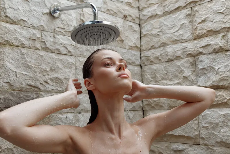 young woman taking shower under rain showerhead with water droplets on skin against stone wall background, relaxation and freshness in bathroom setting