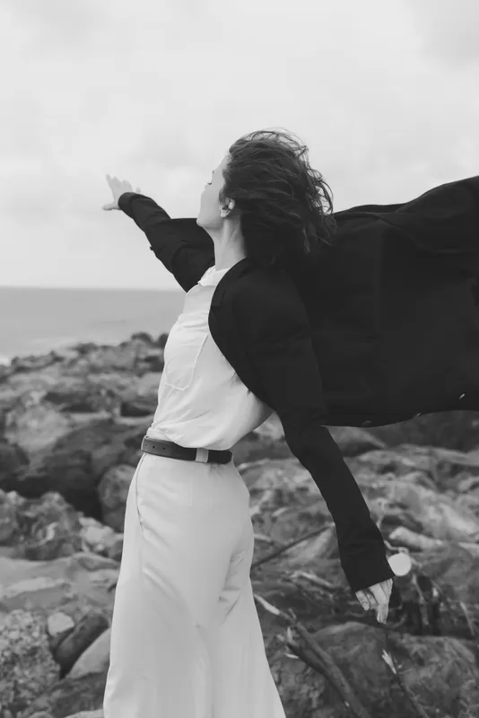 Wind swept cape and dress move along a rugged coastline, embodying freedom, fashion, and dramatic outdoor elegance in monochrome and motion artistry