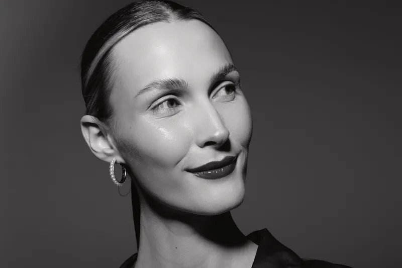 Portrait of a young white woman with light skin and slicked back hair smiling softly, wearing hoop earrings and dark clothing, on a grey background. Concept of elegance and natural beauty.