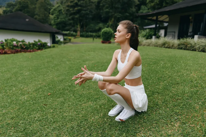 Athletic woman in sporty outfit squatting on green grass in a serene outdoor setting, expressing focus and determination during a fitness routine