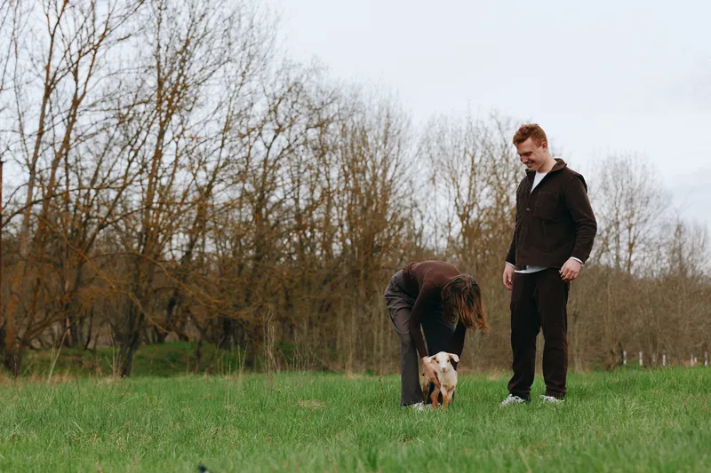 Natural outdoor scene featuring a couple with their dog in a park, bending to interact with the pet, enjoying a calm moment on green grass, bright sky, casual lifestyle, candid companionship, shared
