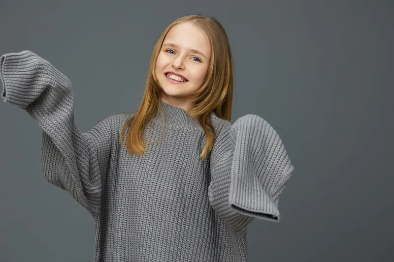 Cheerful girl in oversized grey knitted sweater against minimalistic background, showcasing joy and warmth with casual autumn style