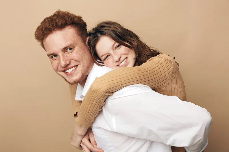 Smiling young couple in casual clothes embraces happily against beige background. The woman hugs the man from behind, both showing joyful expressions. Concept of love, friendship, and togetherness.