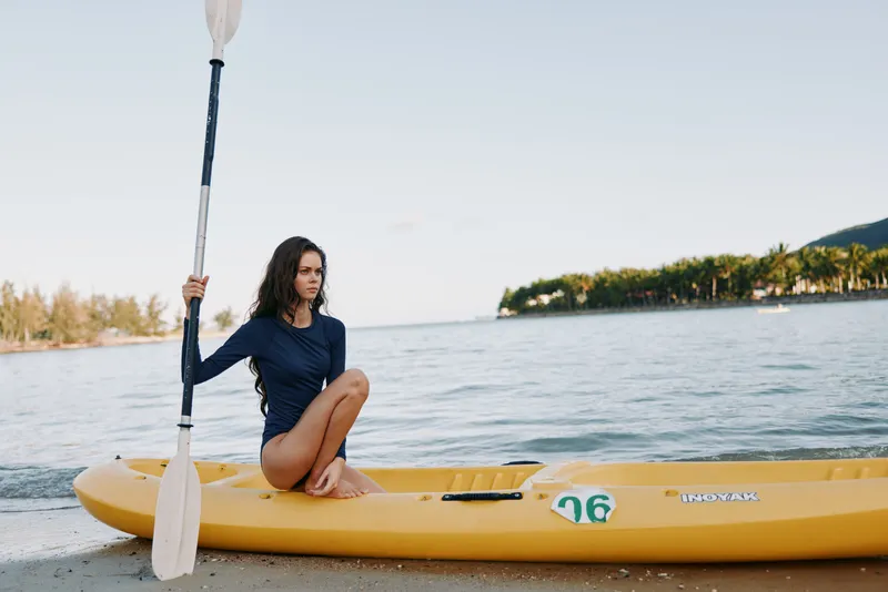 Female kayaker sits on a bright yellow kayak at the shore, barefoot, while holding a paddle. Calm water, clear sky, and serene beach atmosphere convey outdoor adventure.