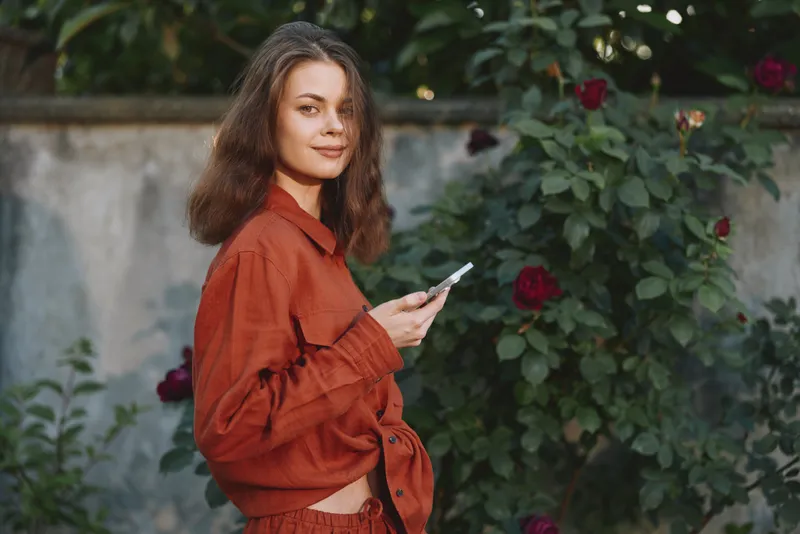 Smiling woman with wavy hair wearing an orange shirt standing in a garden, looking at a smartphone. Casual lifestyle, outdoor ambiance, natural light, everyday moments