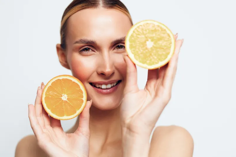 Young woman with blonde hair smiles while holding two halves of a fresh orange fruit near her face against a white background, conveying freshness and health.
