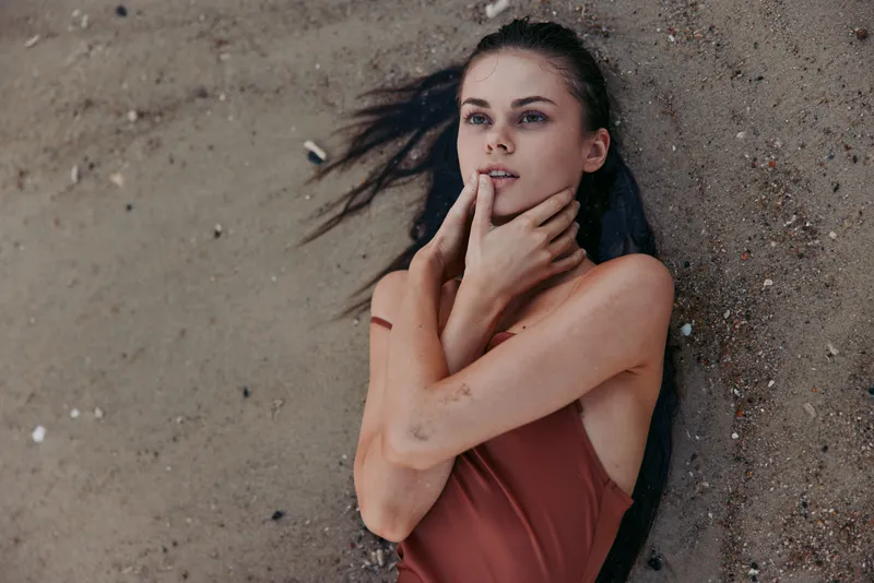 Thoughtful woman in rust swimsuit lying on sandy beach with dark hair spread, expressing calmness and contemplation by the shore.