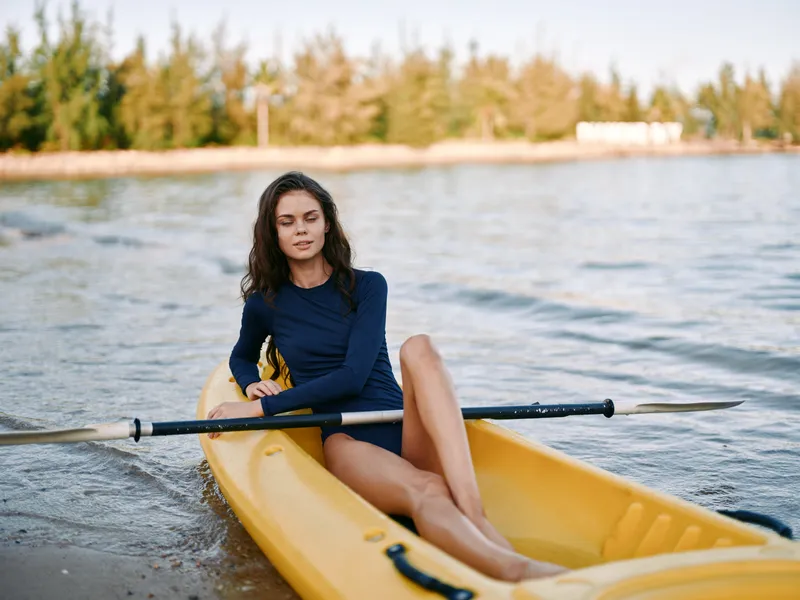 A woman sits calmly in a yellow kayak on calm lake water, wearing a navy long sleeve top and shorts, legs extended as the paddle rests beside her.