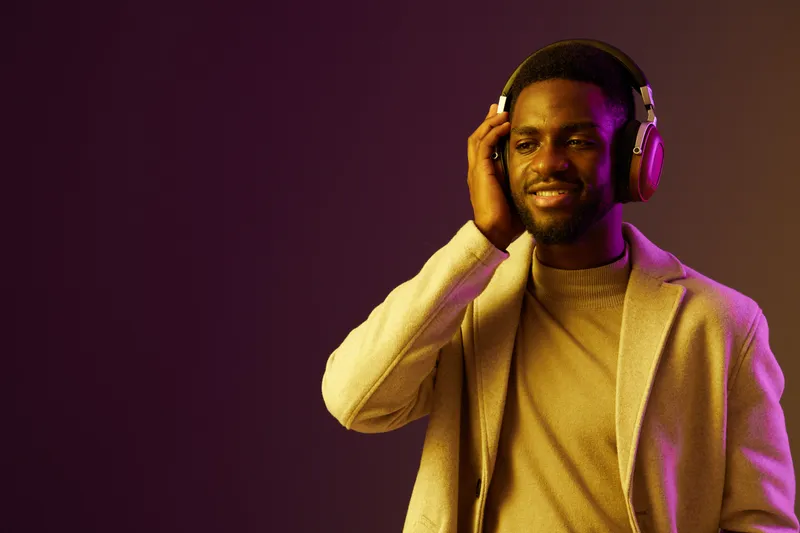 Young African man enjoying music with headphones in stylish beige blazer, smiling against a gradient purple background, conveying happiness and relaxation
