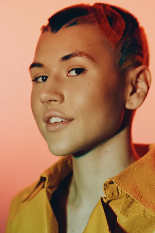 Close-up portrait of a confident woman with short red hair and natural makeup wearing a yellow textured shirt against a warm peach background.
